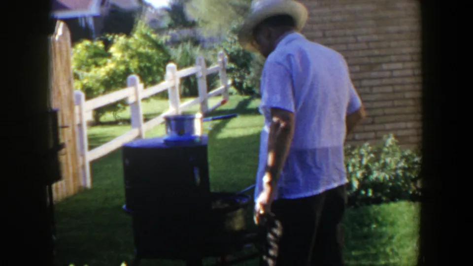 The 1953 home-movie frame shows the man beside a dark grill, with a pot, white fence, lawn, and brick wall in the yard.