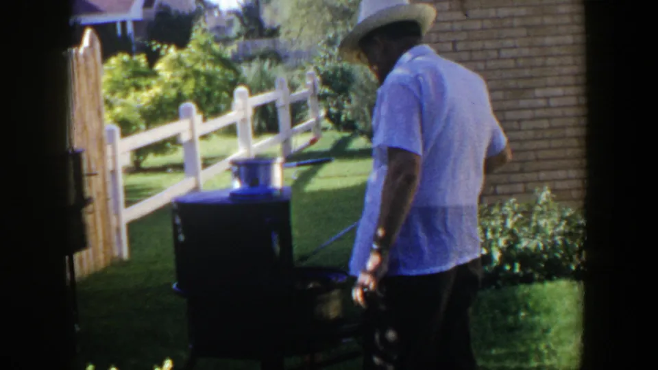 A man in a white fedora stands at the grill in the Lubbock backyard, framed by lawn, fence rails, plants, and a brick wall.
