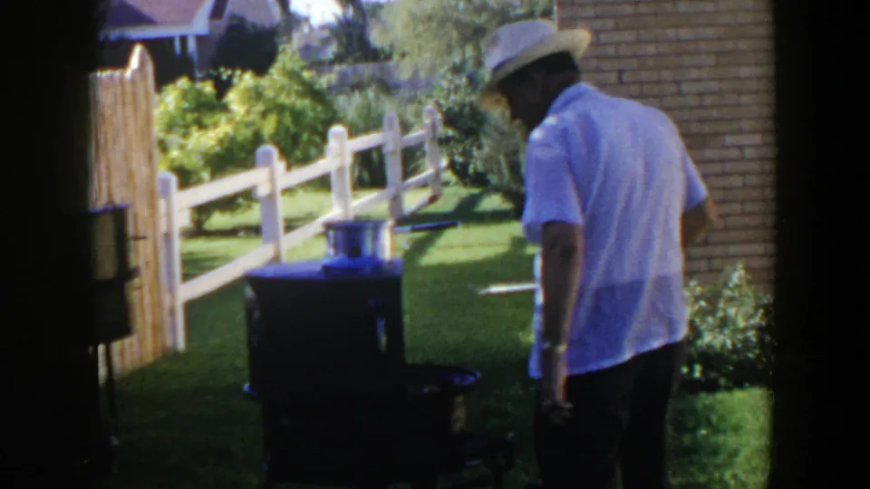 The man moves beside the grill in a sunny yard, with a white fence, lawn, shrubs, and brick wall visible behind him.