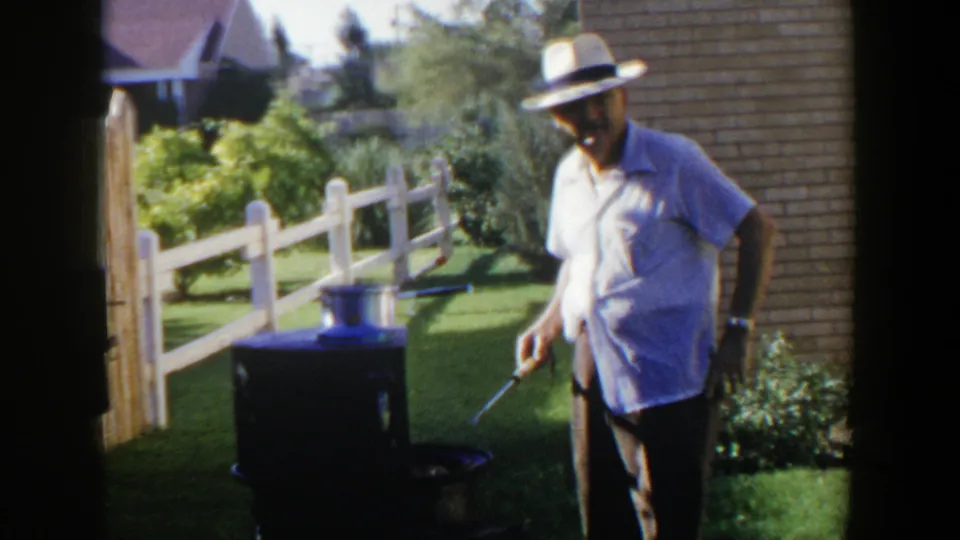 The man in a straw hat and short-sleeve shirt faces the camera while holding a tool near the outdoor grill in the yard.