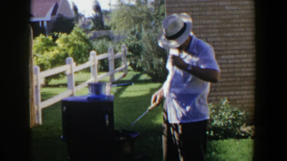The man looks down near the grill, one hand raised near his straw hat, with the backyard fence and brick wall behind him.