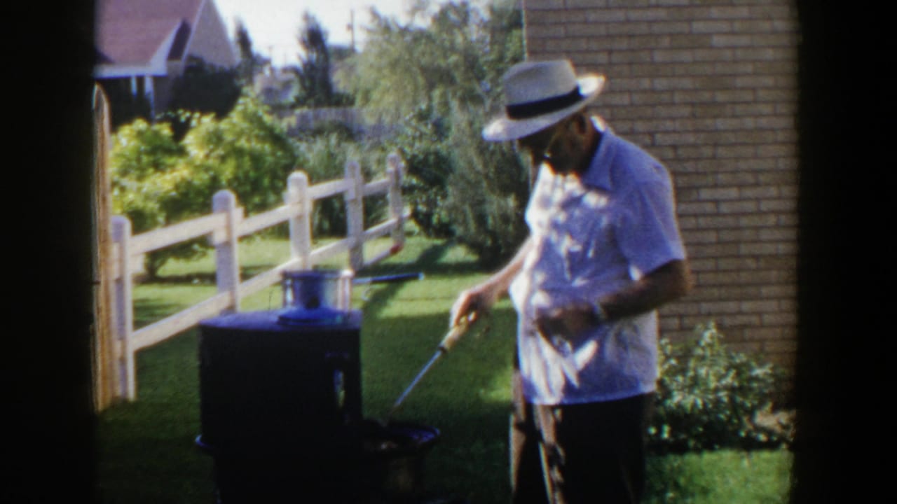 Older man in a white hat tends a small outdoor cooker in a sunny yard.