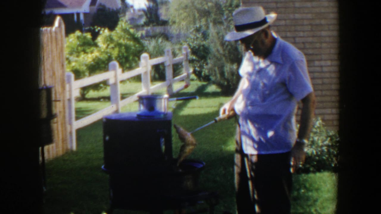 Man in a white hat uses a long utensil near a small outdoor grill.