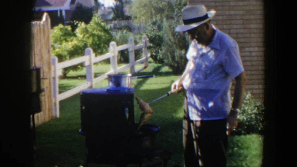 An older man in a white hat uses a long-handled utensil while cooking outdoors in a Lubbock backyard.