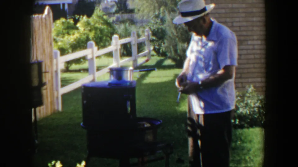The man pauses beside the outdoor cooker, with a pot on top and a fenced green yard behind him.