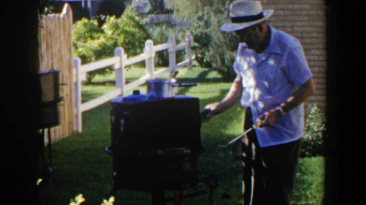 In 1953 Lubbock backyard footage, a man in a light shirt and fedora tends a dark outdoor grill near a white fence.