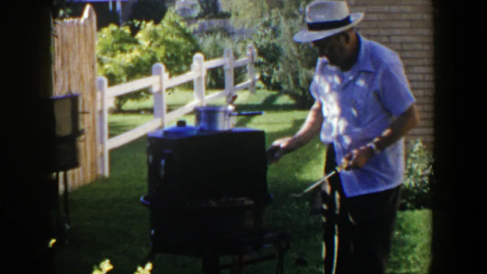 In 1953 Lubbock backyard footage, a man in a light shirt and fedora tends a dark outdoor grill near a white fence.