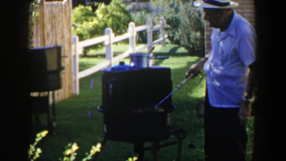 A man in a white hat works at a black grill in a sunny Lubbock, Texas yard, with a pot visible on top.