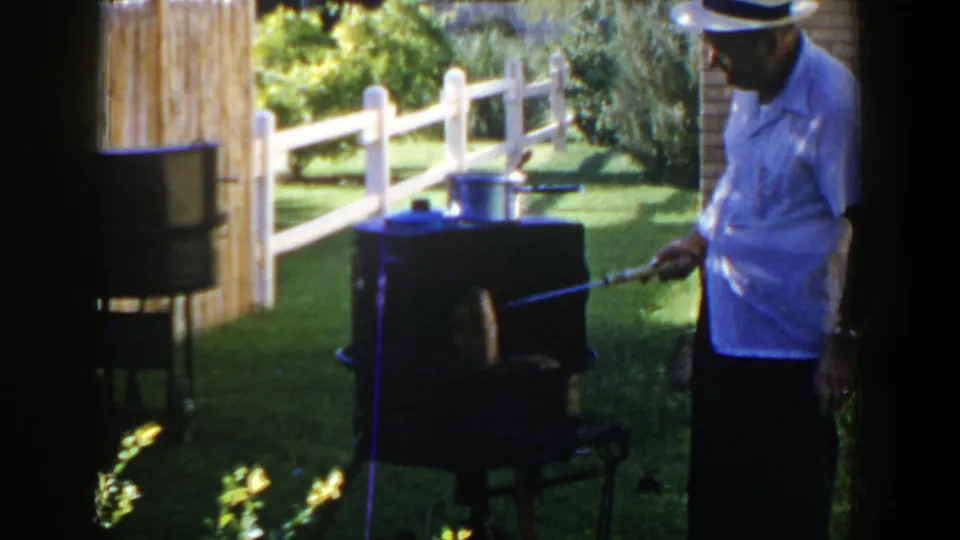The 8mm frame shows the man close to the backyard grill, holding a tool near the front opening in bright sunlight.