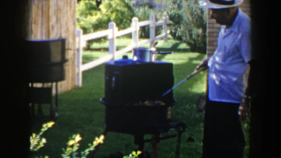 A man in a hat stands beside the dark outdoor grill in the 1953 Lubbock yard, with fencing and grass behind him.