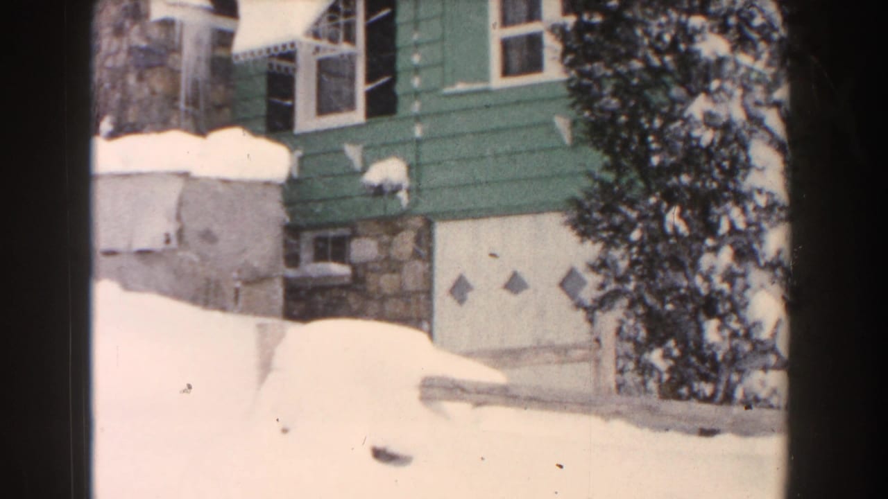 Snow-covered green bungalow with stonework, garage door, awnings, and deep snow in the yard.