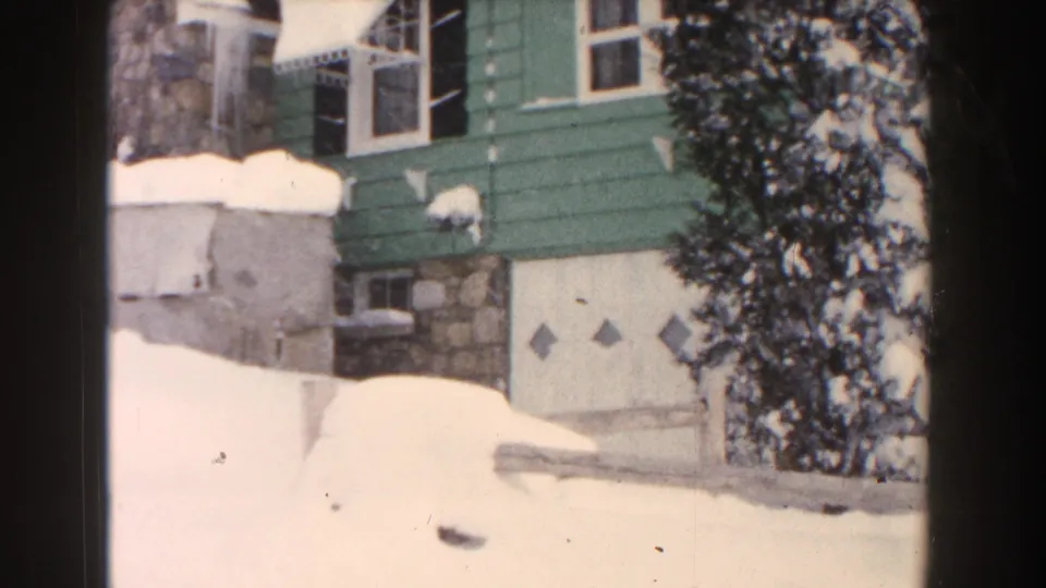 1961 Dearborn, Michigan home-movie frame showing a green-sided bungalow with stone accents and heavy snow piled around the garage and roof.