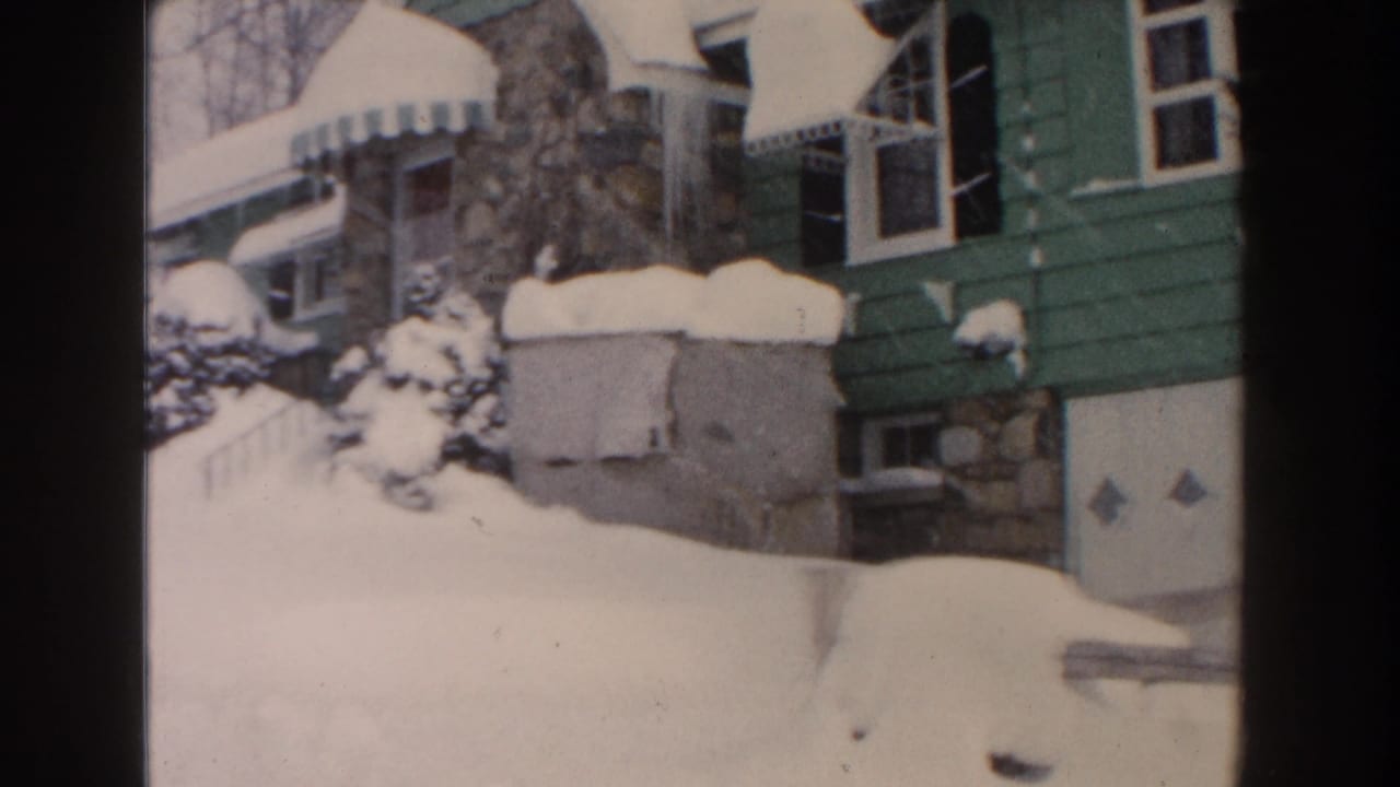 Snow piled around a house entrance, garage, awning, shrubs, and steep roofline.