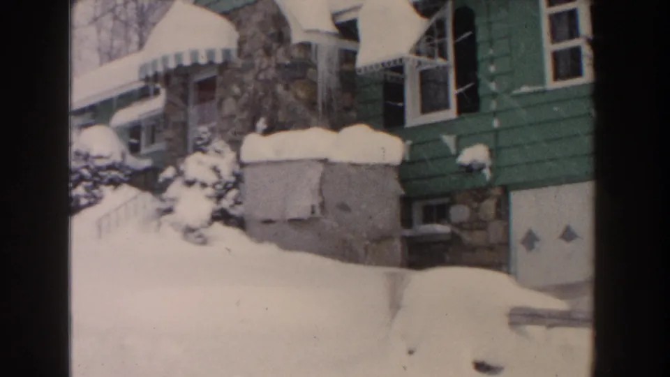 1961 Dearborn, Michigan frame showing the bungalow from a slightly shifted angle, with snow heaped on the roof, shrubs, and garage area.