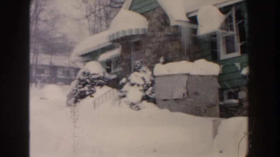 1961 Dearborn, Michigan home-movie frame with the camera panned left, revealing snow-covered shrubs and a neighboring building beyond the bungalow.
