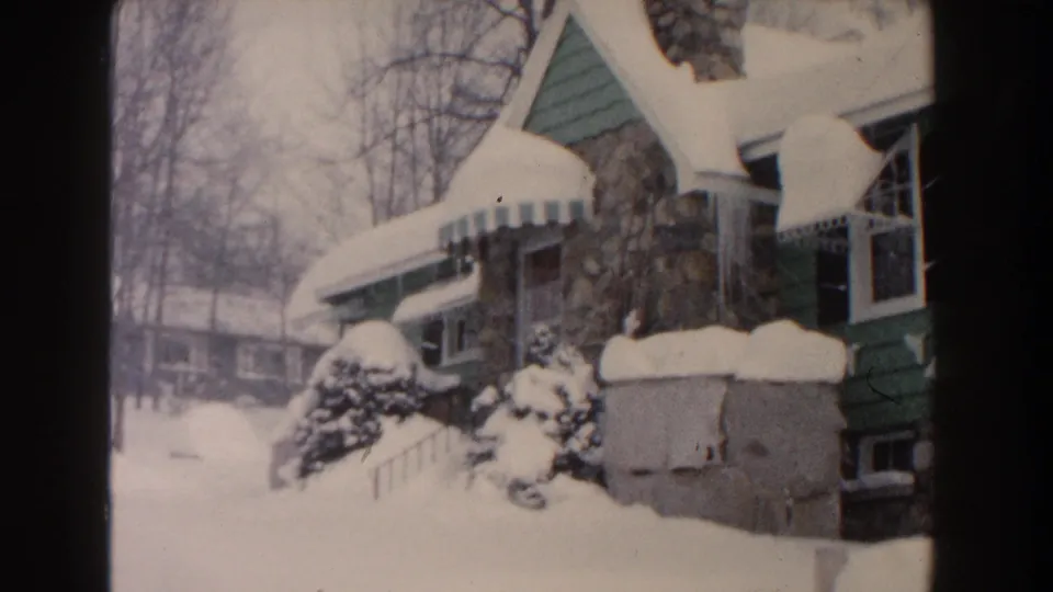 1961 Dearborn, Michigan frame showing the side of the snow-covered bungalow with a background building and bare winter trees.