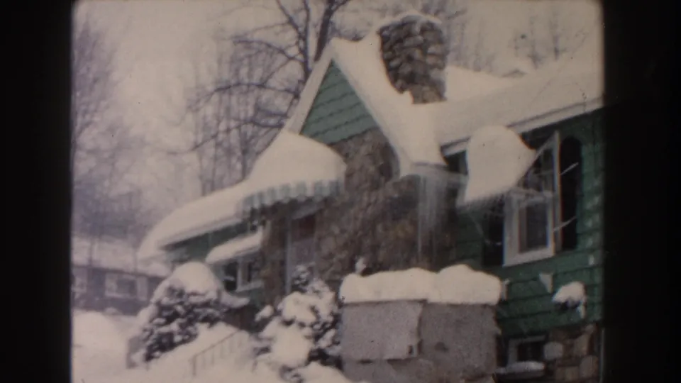 1961 Dearborn home-movie frame showing a green house under heavy snow, with a stone chimney, icicles, awning, shrubs, and bare winter trees.