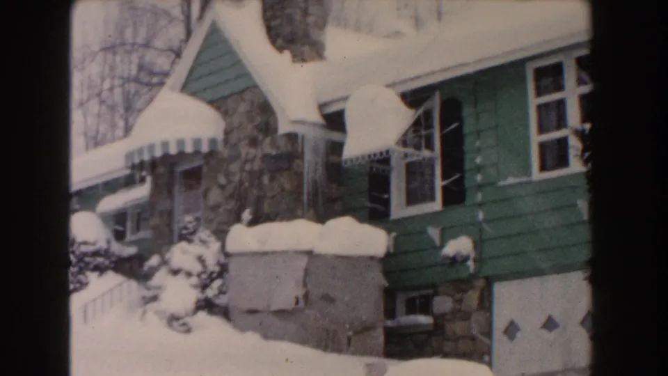 1961 Dearborn home-movie view of a green house partly buried in snow, with stonework, awning, shrubs, basement window, and garage door visible.