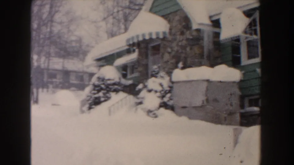 1961 Dearborn frame showing the snow-covered yard and green house exterior, with a stone chimney, awning, shrubs, and bare trees in soft 8mm color.