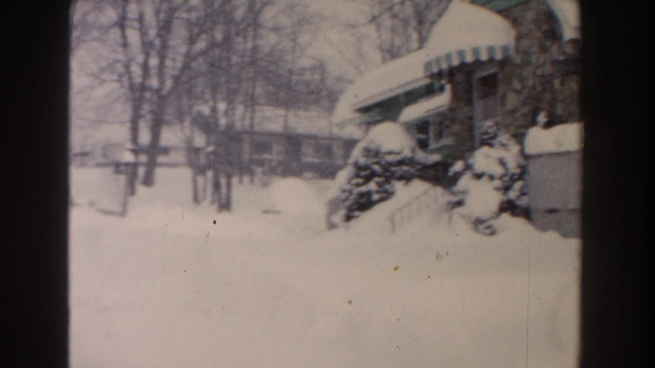 Snow-covered residential street with house, awning, shrubs, and bare trees.