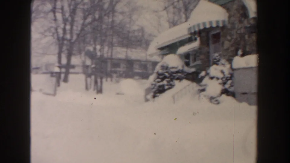 1961 Dearborn home-movie frame showing a snow-covered residential street, a house with striped awning, shrubs, and leafless trees.