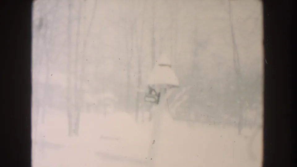 Soft 8mm frame from Dearborn showing a snow-laden mailbox or post against bare winter trees and a washed, snowy background.