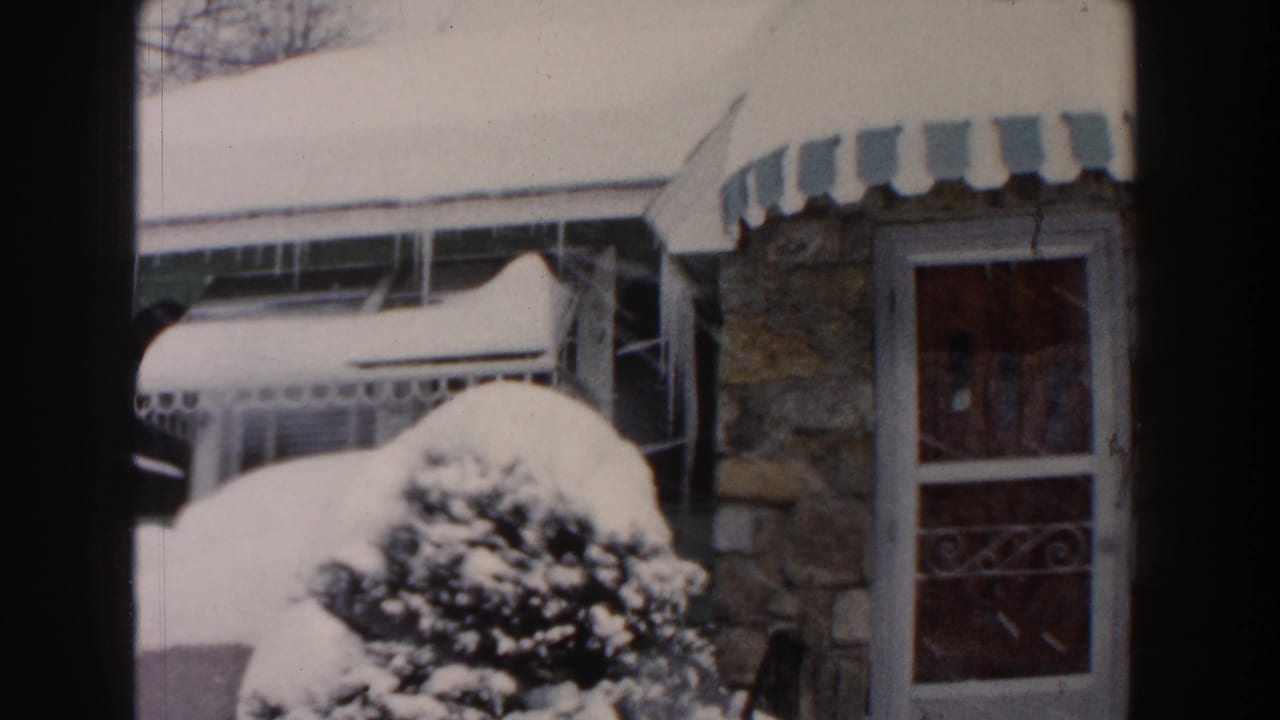 1961 Dearborn home-movie frame showing a snow-covered stone bungalow entrance with a striped awning and evergreen shrub.
