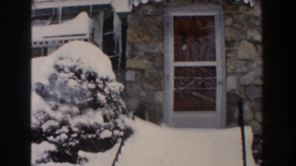 1961 Dearborn exterior frame with a stone house wall, front door, deep snow, and shrubs weighed down by snowfall.