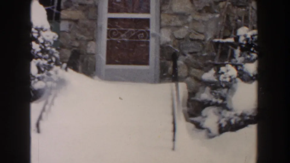 1961 Dearborn frame showing the lower front entrance, snow-covered walkway, stone facade, and a shrub beside the door.