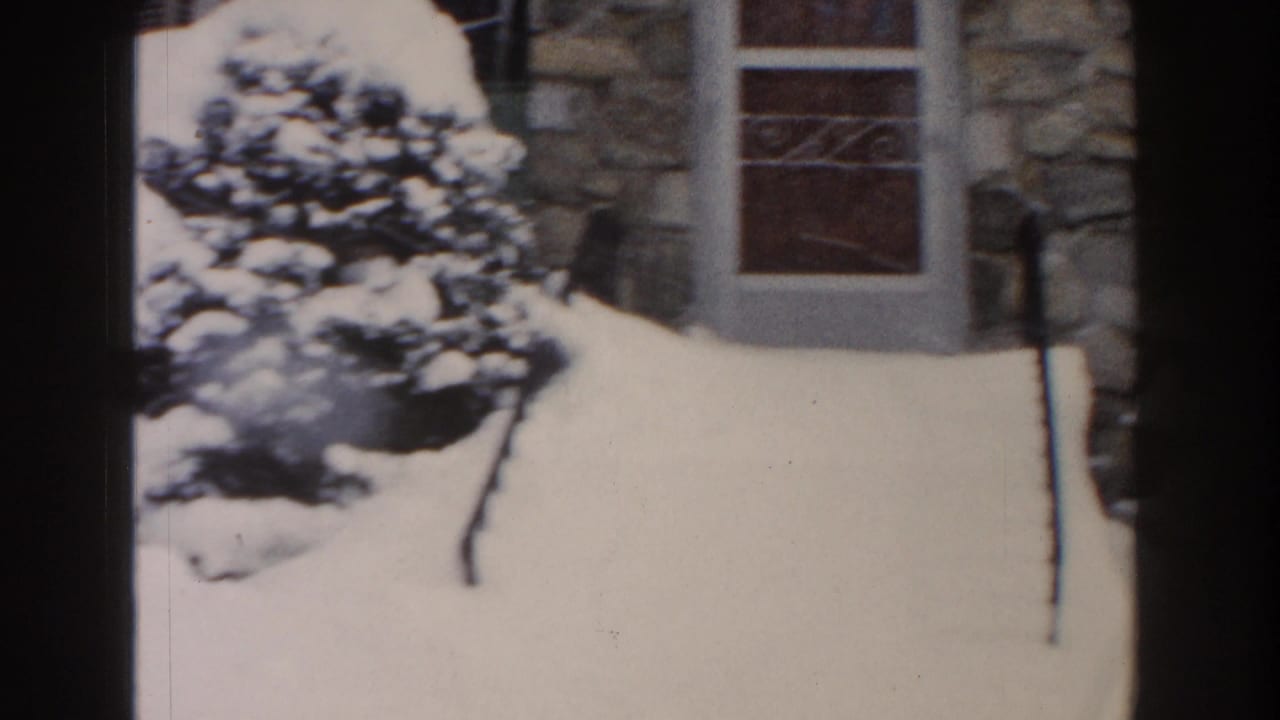 1961 Dearborn home-movie frame showing deep snow around an evergreen beside a stone residential facade and window.