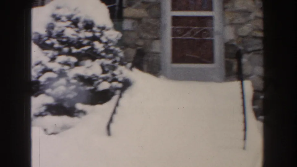 1961 Dearborn home-movie frame showing deep snow around an evergreen beside a stone residential facade and window.