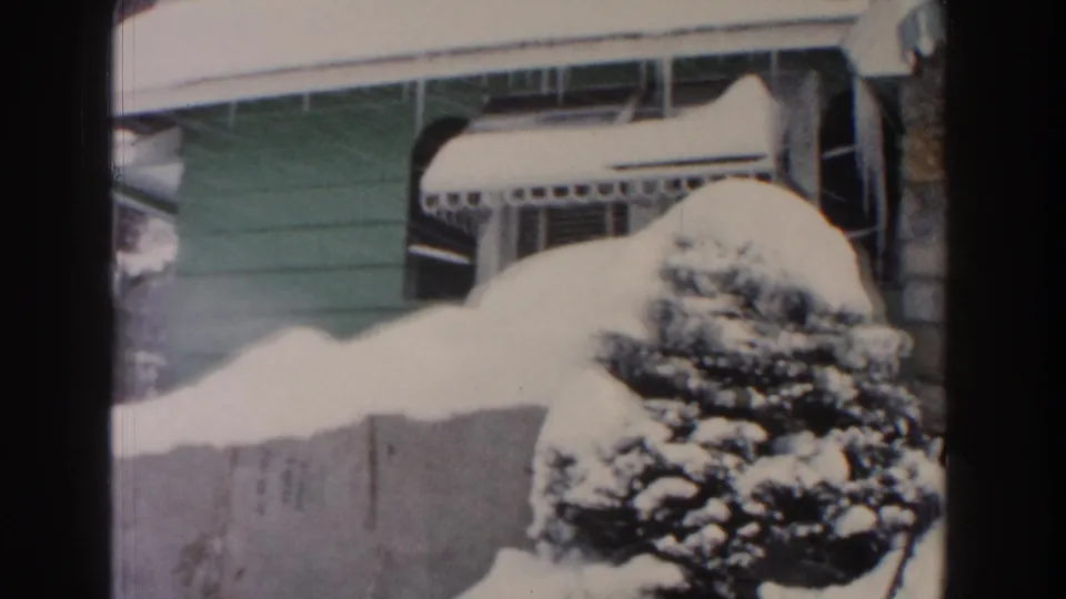1961 Dearborn 8mm frame showing a green-sided bungalow exterior, striped porch awning, evergreen, and deep accumulated snow.