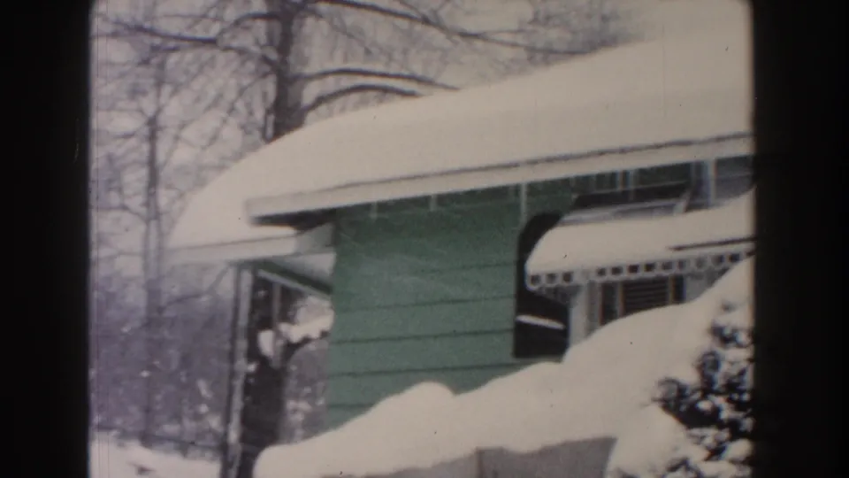 1961 Dearborn frame showing a green bungalow exterior, snow-covered roofline, striped awning, bare trees, and piled snow.