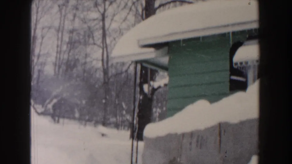 1961 Dearborn home-movie frame showing a green-sided house, heavy snow on the roof, and bare winter trees.