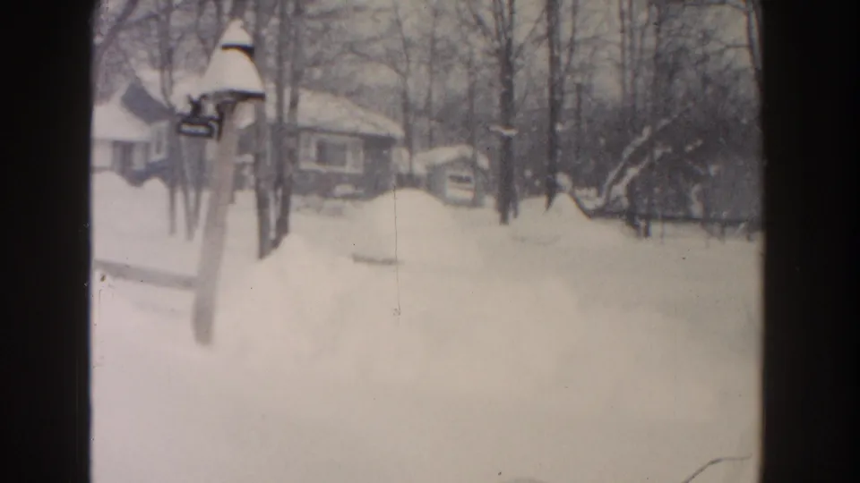 1961 Dearborn frame looking across a snow-filled yard toward bare trees, low houses, and a small shed-like structure.
