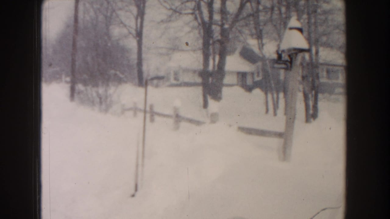 1961 Dearborn, Michigan home-movie frame showing a snowy residential roadside with bare trees, small houses, fencing, and a bird feeder.