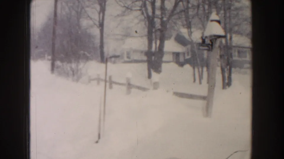 1961 Dearborn, Michigan home-movie frame showing a snowy residential roadside with bare trees, small houses, fencing, and a bird feeder.