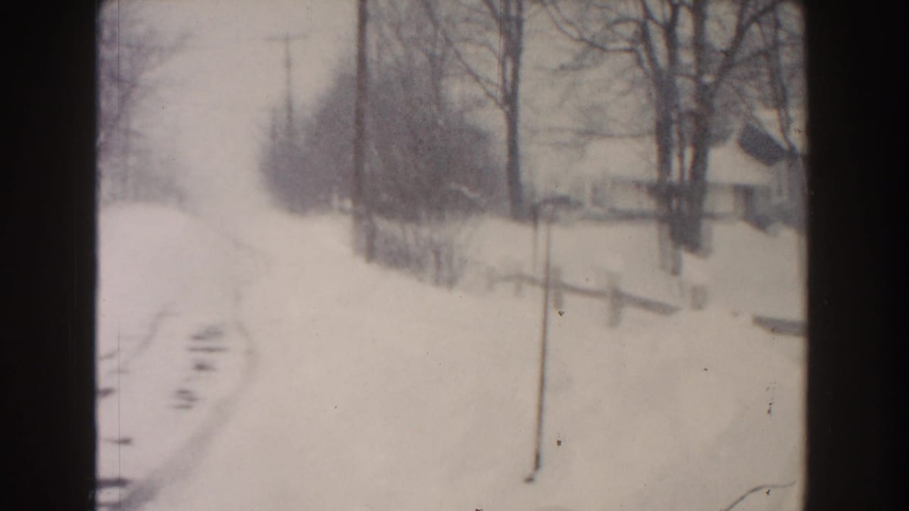 1961 Dearborn, Michigan winter frame with a snow-covered road, tire tracks, bare trees, utility poles, fencing, and a small house.