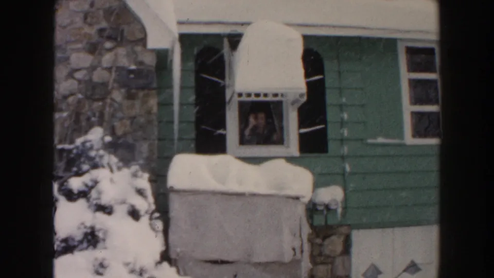 1961 Dearborn home-movie frame showing a person at a window of a green-sided house during snowfall.