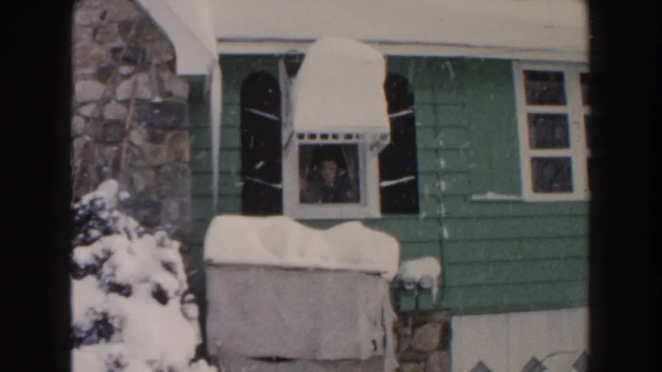 Vintage 8mm frame of a Dearborn house exterior with a person at the window during winter snowfall.
