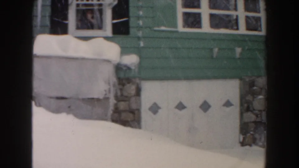 Lower portion of the 1961 Dearborn bungalow showing green siding, icicles, stone supports, and a white garage door in snow.
