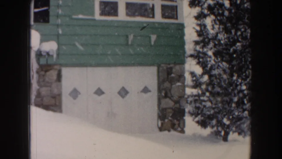 Dearborn winter home-movie frame with the bungalow garage, green siding, stone trim, icicles, and a snow-dusted evergreen.