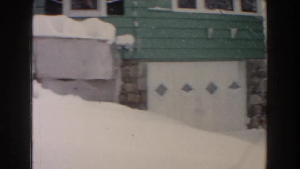 1961 Dearborn home-movie frame showing a green house, stonework, white garage door, and deep snow in the yard.