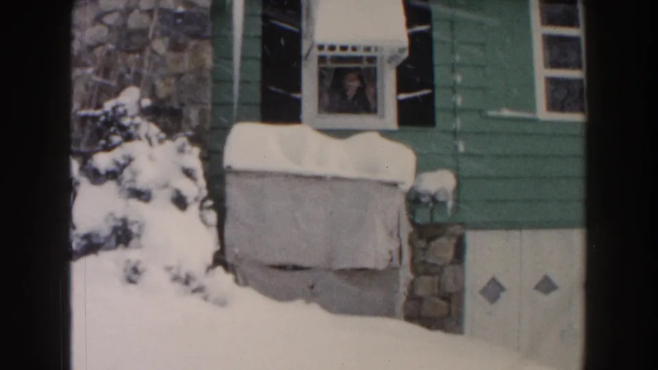 1961 Dearborn frame of a snow-covered green house with black shutters and a person visible behind an upstairs window.