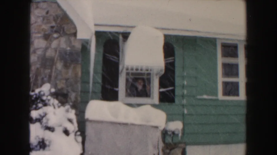 1961 Dearborn home-movie frame showing a person at an upstairs window, black shutters, stonework, and heavy snow on an awning.