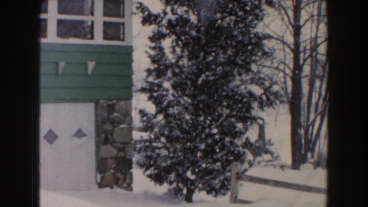 1961 Dearborn, Michigan 8mm frame showing a snow-covered residential exterior with green siding, white upper walls, multi-pane windows, evergreen trees, and bare winter branches.