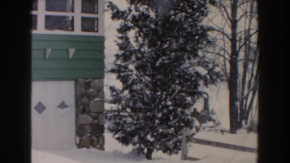 1961 Dearborn, Michigan 8mm frame showing a snow-covered residential exterior with green siding, white upper walls, multi-pane windows, evergreen trees, and bare winter branches.