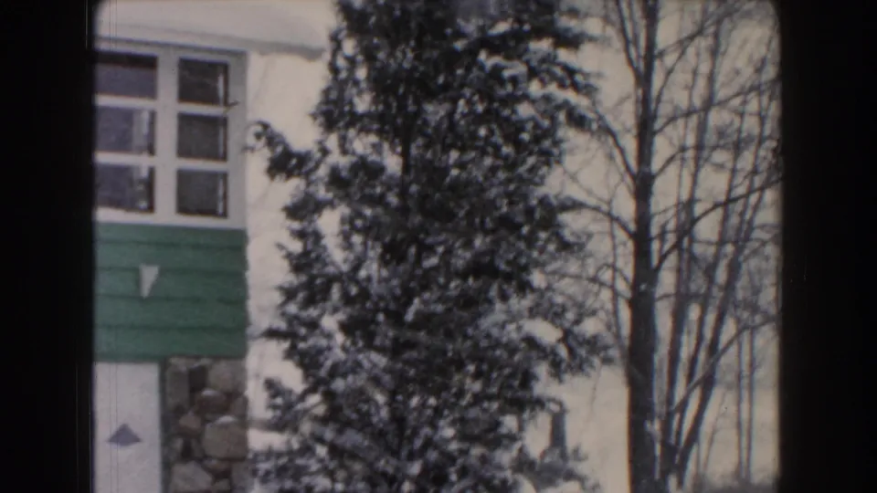 1961 Dearborn, Michigan home-movie frame with a green-and-white residential facade, snow-covered evergreen trees, bare branches, and dark vertical frame edges from the scanned film.