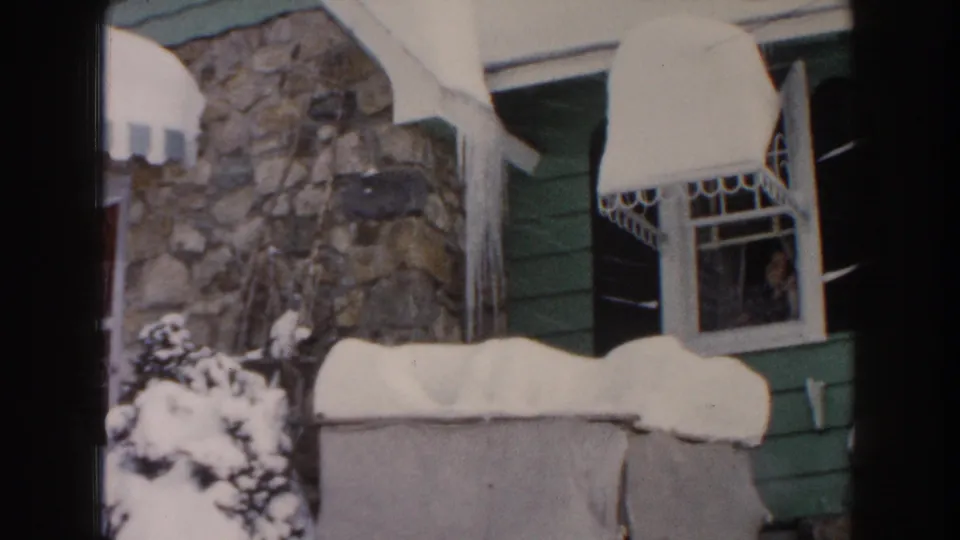 1961 Dearborn, Michigan 8mm frame showing a closer residential detail with stonework, green siding, white awnings, roofline snow, icicles, and a window during winter conditions.