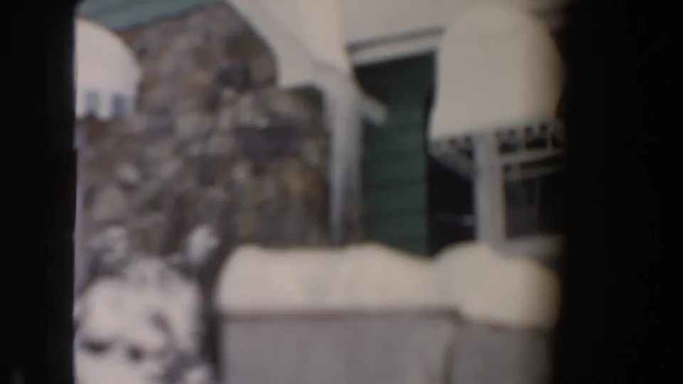 Soft-focus 1961 Dearborn home-movie frame of a snow-covered residential facade with awning and stonework.